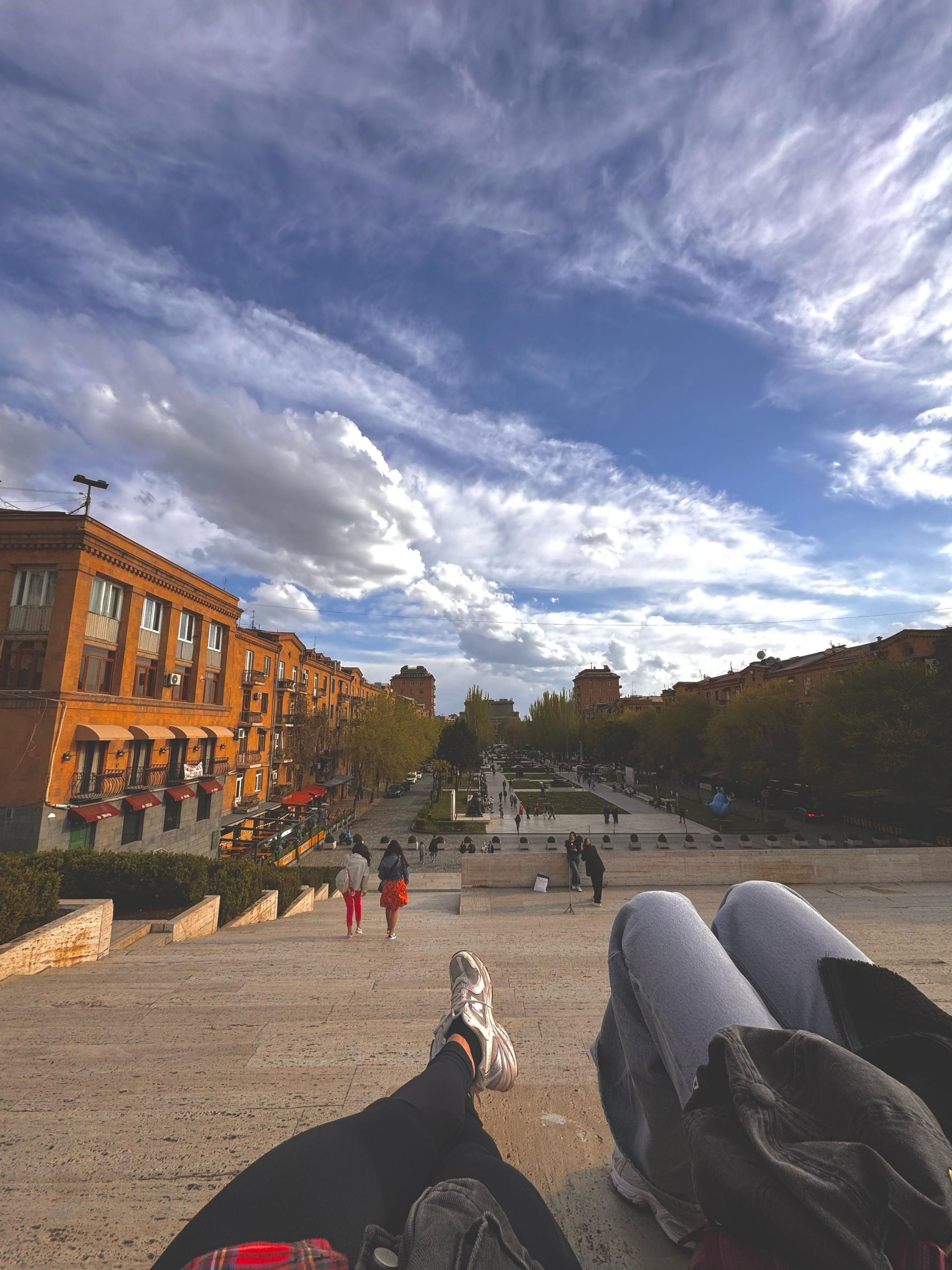 Una vista in prima persona di due persone sedute su un'ampia scalinata in pietra che si affaccia su una piazza cittadina con persone che camminano sotto.
