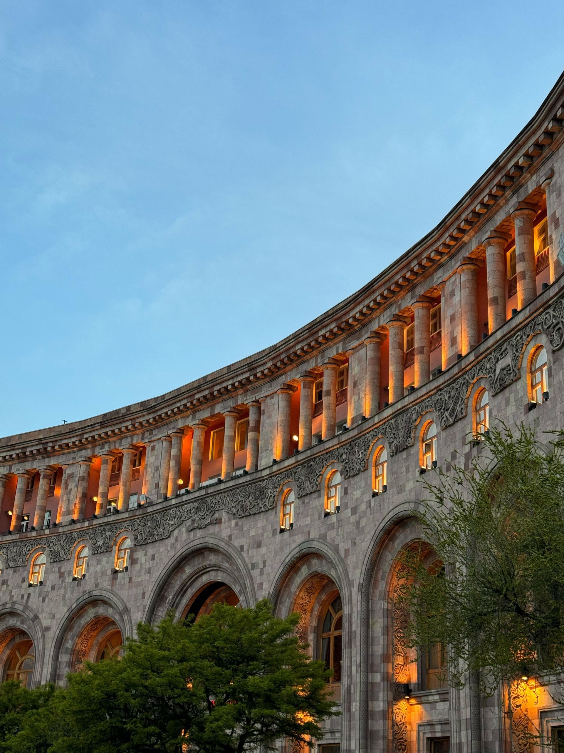 Un grande edificio in pietra ricurvo con colonne e archi classici, illuminato da luci calde contro un cielo crepuscolare.