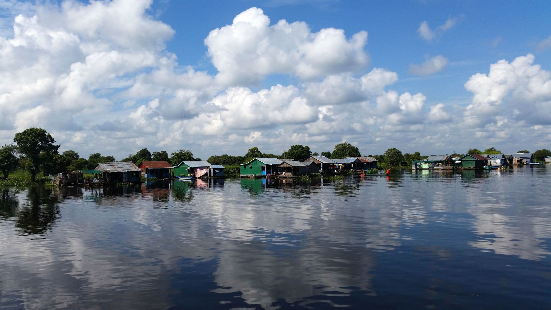 Un village flottant avec des maisons colorées sur une rivière calme, reflétant le ciel bleu et les nuages blancs.