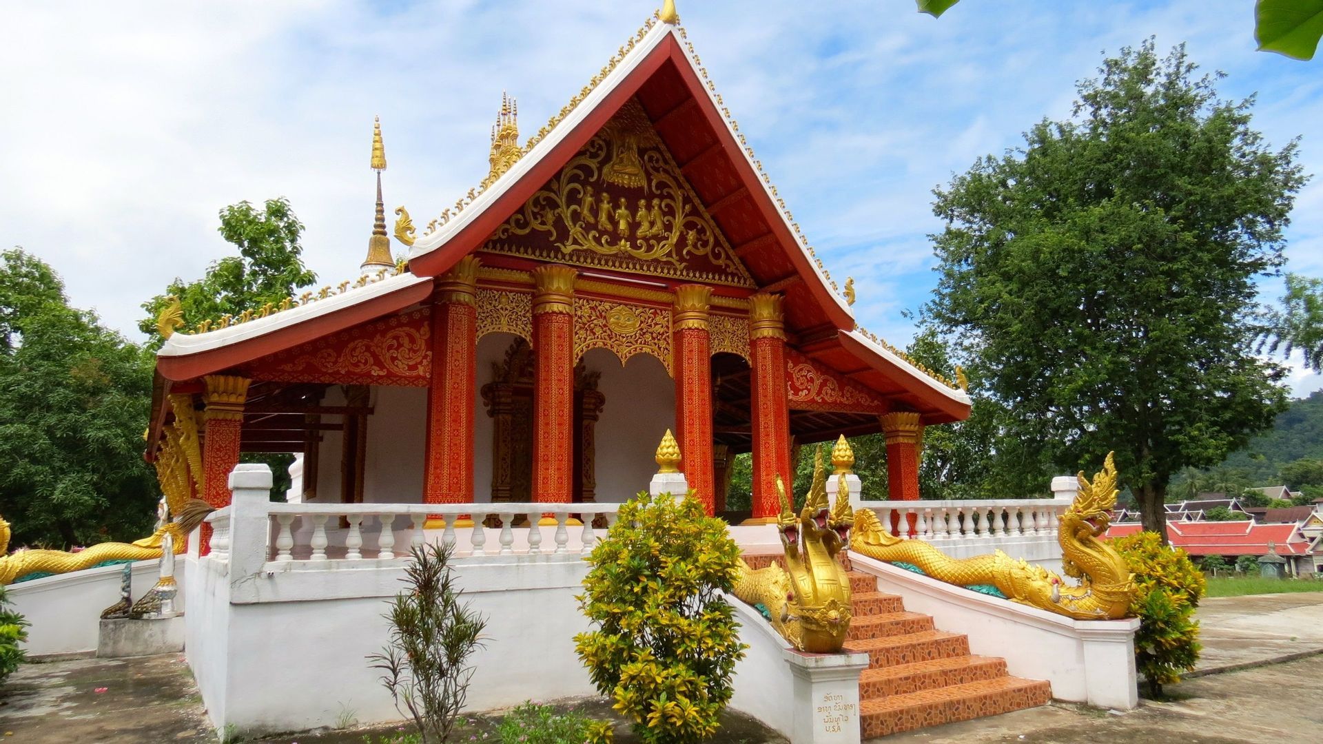 Un temple bouddhiste orné de rouge et d'or, avec des statues de naga dorées le long de son escalier, se dressant sur fond d'arbres et de ciel bleu.
