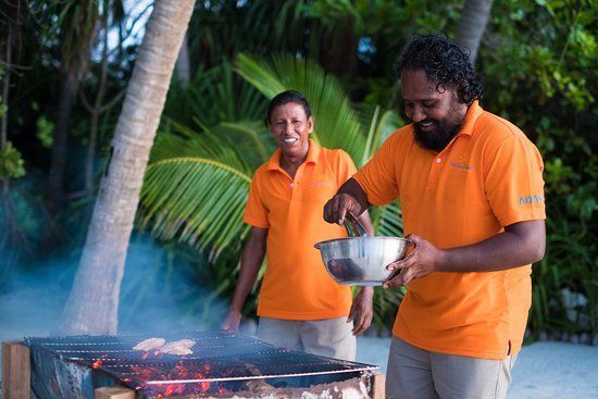 Due uomini con polo arancioni abbinate sorridono mentre preparano cibo su una griglia a carbone all'aperto, con palme sullo sfondo.