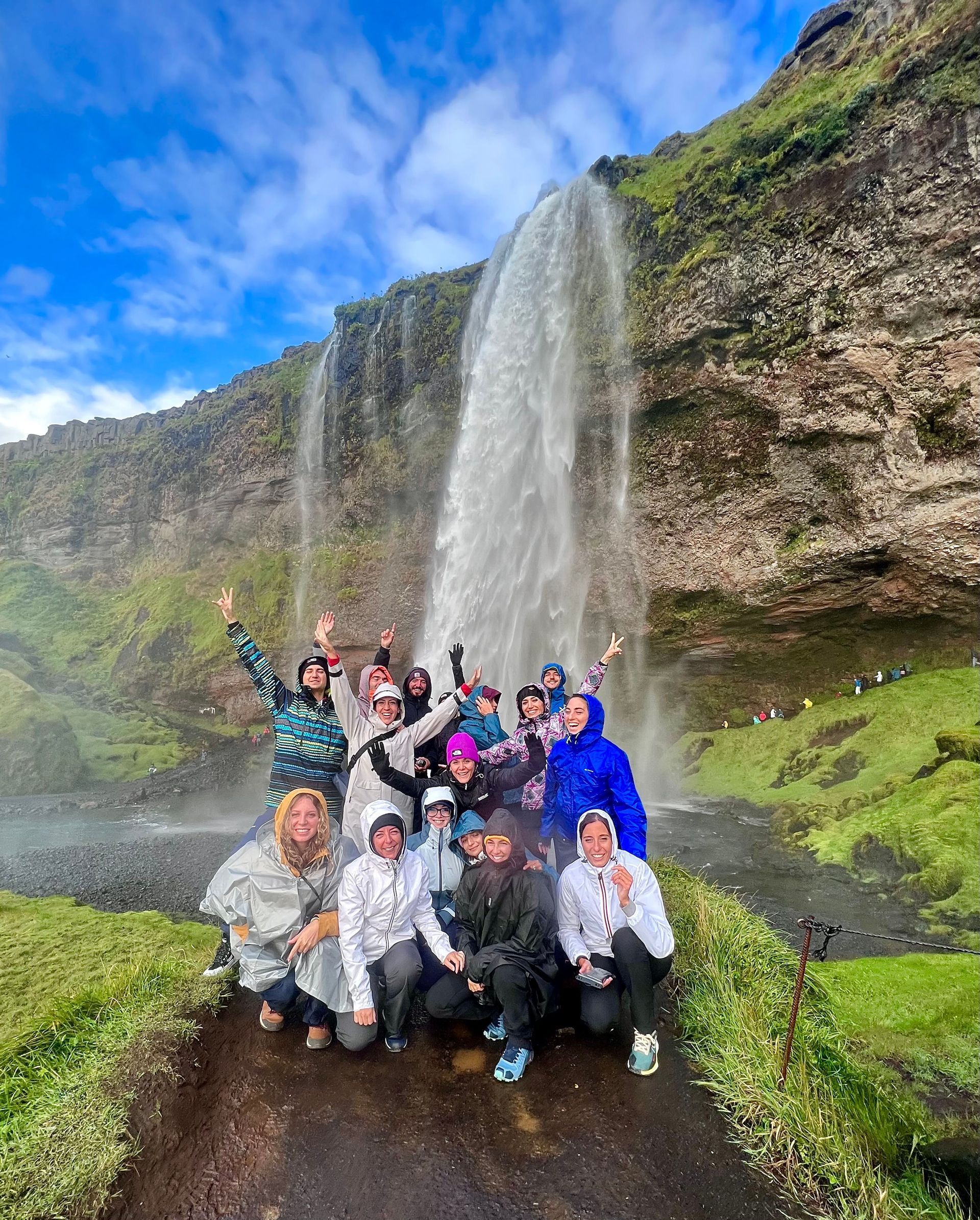 Un groupe WeRoad en imperméables sourit et pose pour une photo devant une grande cascade sur une falaise moussue.