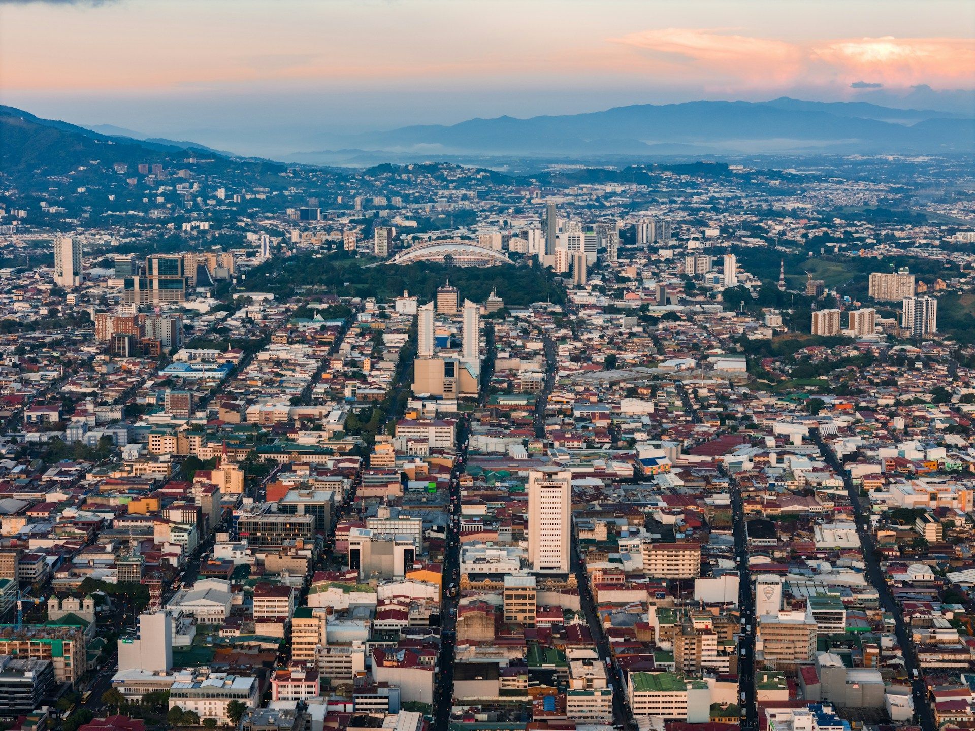 Una vista aérea de un denso paisaje urbano al amanecer, con calles y edificios que conducen hacia las montañas en el horizonte.