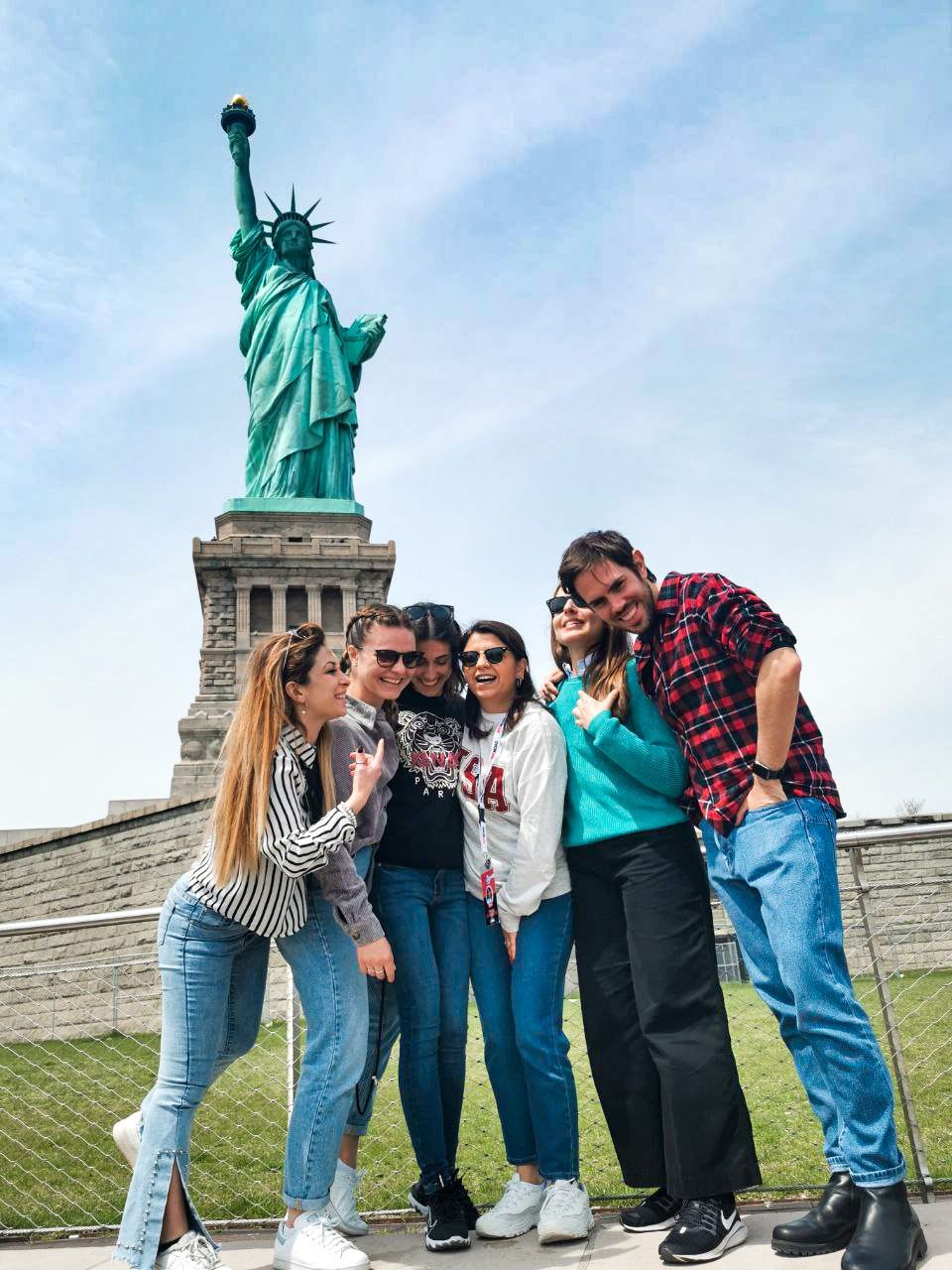 Un gruppo WeRoad sorridente in foto davanti alla Statua della Libertà sotto un cielo sereno.