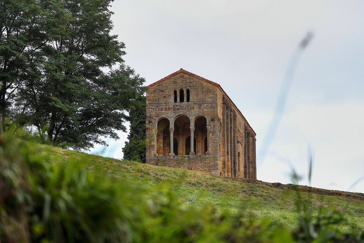 Una antigua iglesia de piedra con fachada arqueada se erige en una colina de hierba junto a un gran árbol bajo un cielo nublado.
