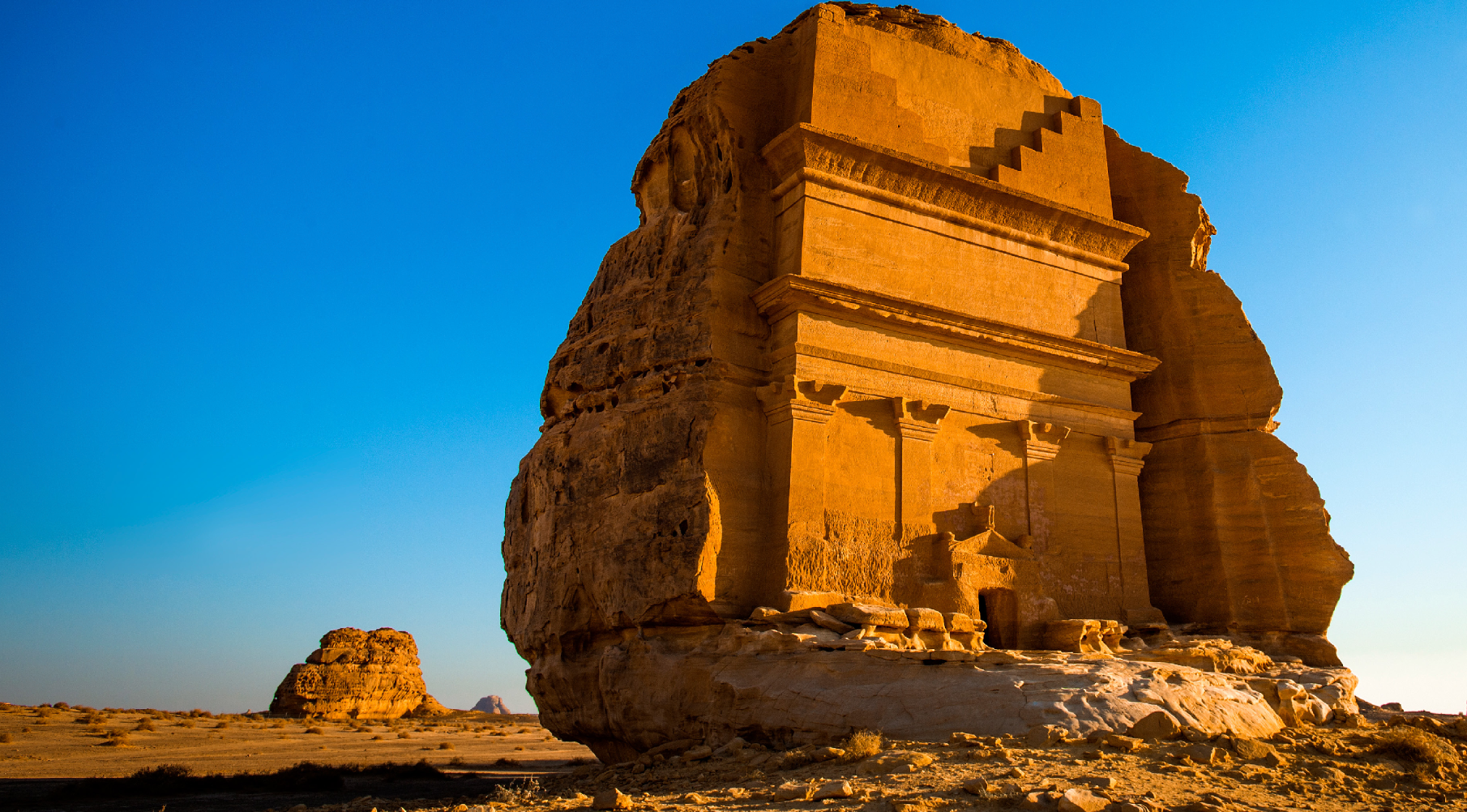 Una tomba monumentale scavata nella roccia arenaria risplende sotto il sole dorato in un vasto deserto sotto un cielo azzurro e limpido.