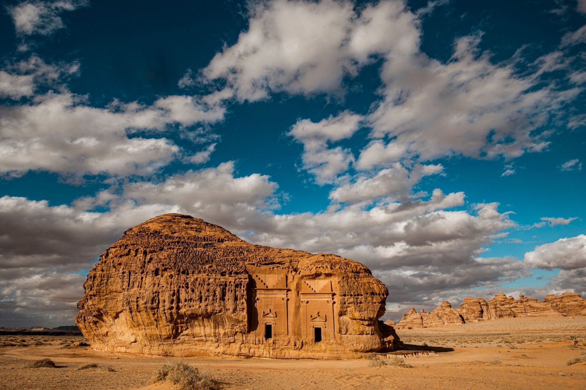 Un'antica tomba scavata in un grande monolite di arenaria nel deserto sotto un cielo azzurro brillante con nuvole bianche.