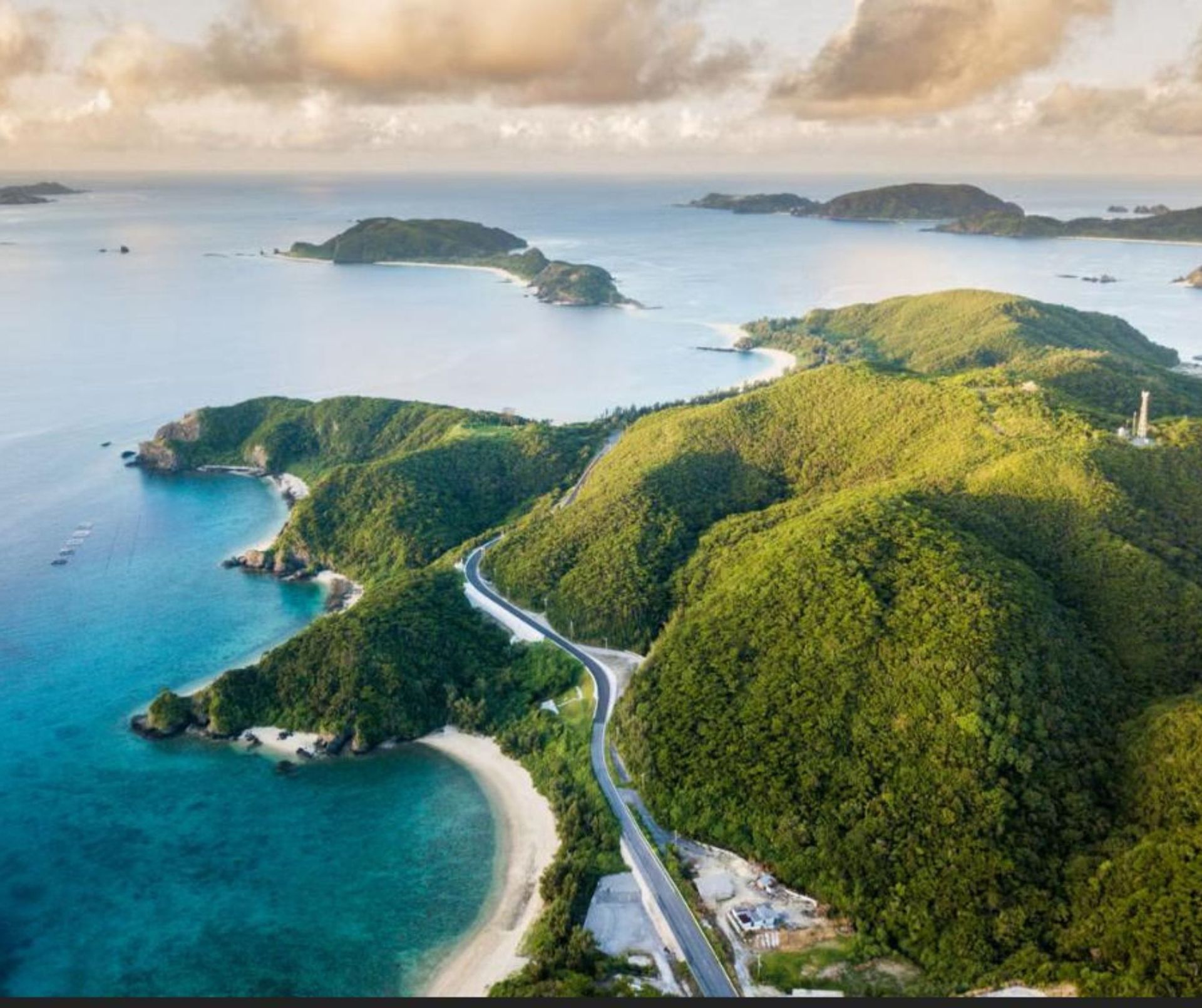Vista aérea de una carretera sinuosa a lo largo de una costa de colinas verdes, con pequeñas islas, calas de arena y agua turquesa.