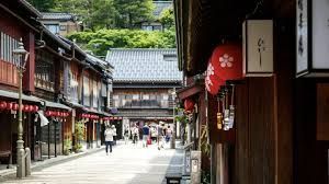 Vista de una calle de un pueblo japonés tradicional con gente caminando entre edificios de madera oscura, con una linterna de papel roja en primer plano.