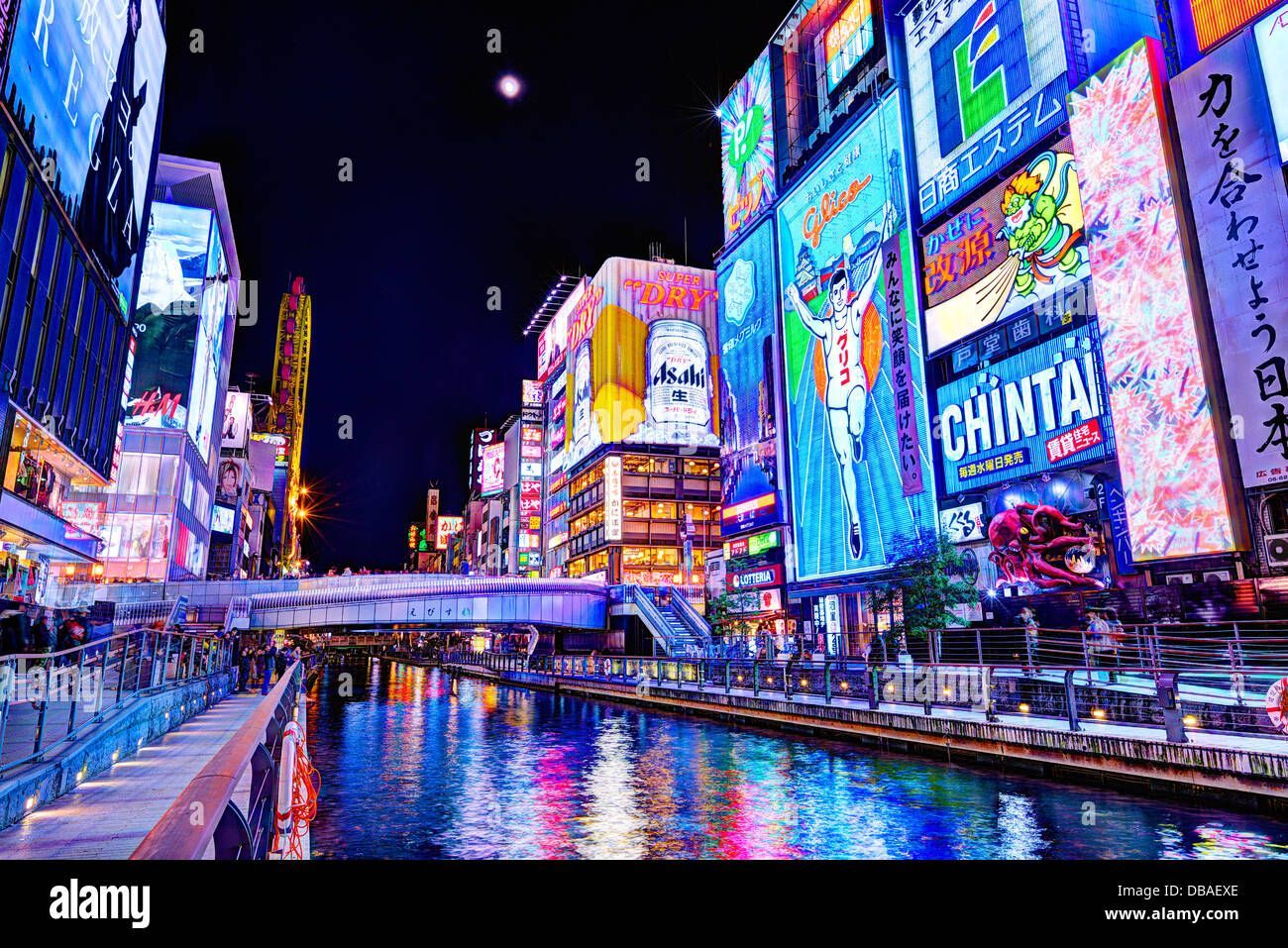 Enormes y coloridos letreros de neón en edificios de la ciudad iluminan un ancho río por la noche mientras la gente pasea por un malecón.