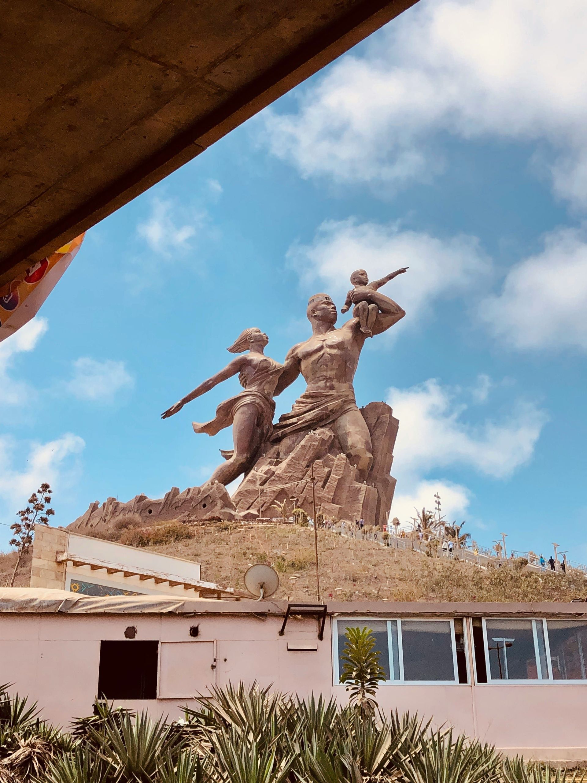 Une statue monumentale en bronze représentant une famille se dresse au sommet d'une colline rocheuse sous un ciel bleu parsemé de nuages blancs et cotonneux.