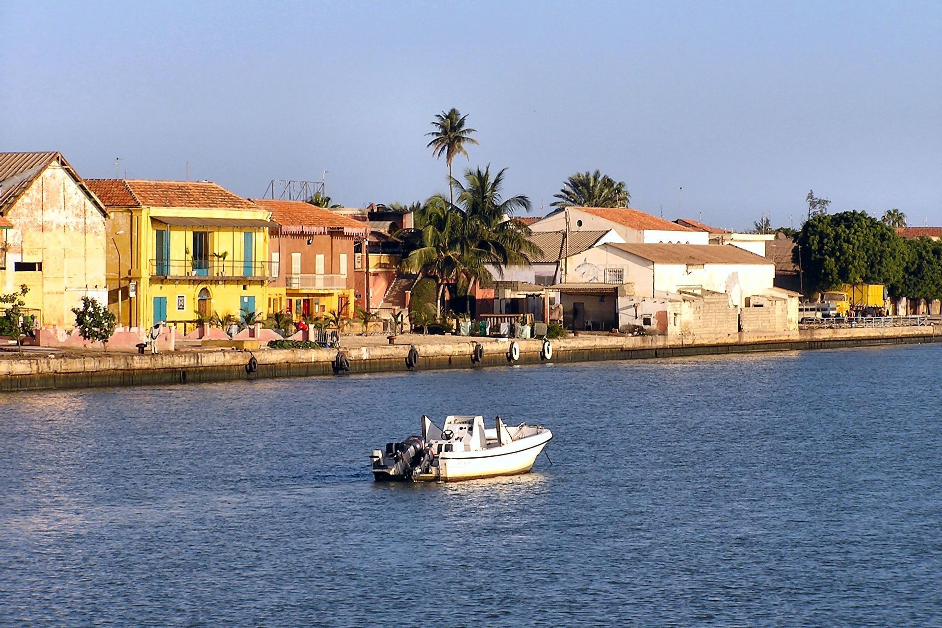 Un bateau à moteur blanc sur l'eau, devant des bâtiments colorés en bord de mer et des palmiers sous un ciel dégagé.