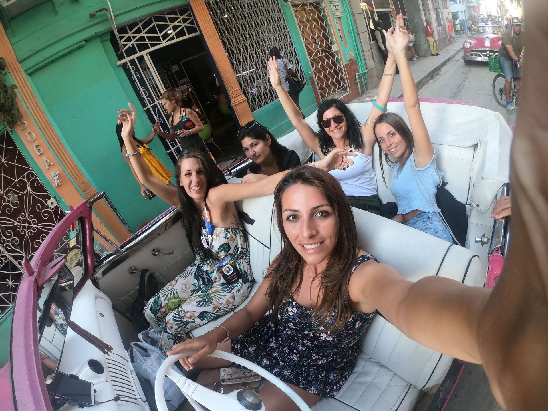 Un gruppo WeRoad di donne si scatta un selfie sorridente a bordo di un'auto decappottabile classica rosa, per strada.