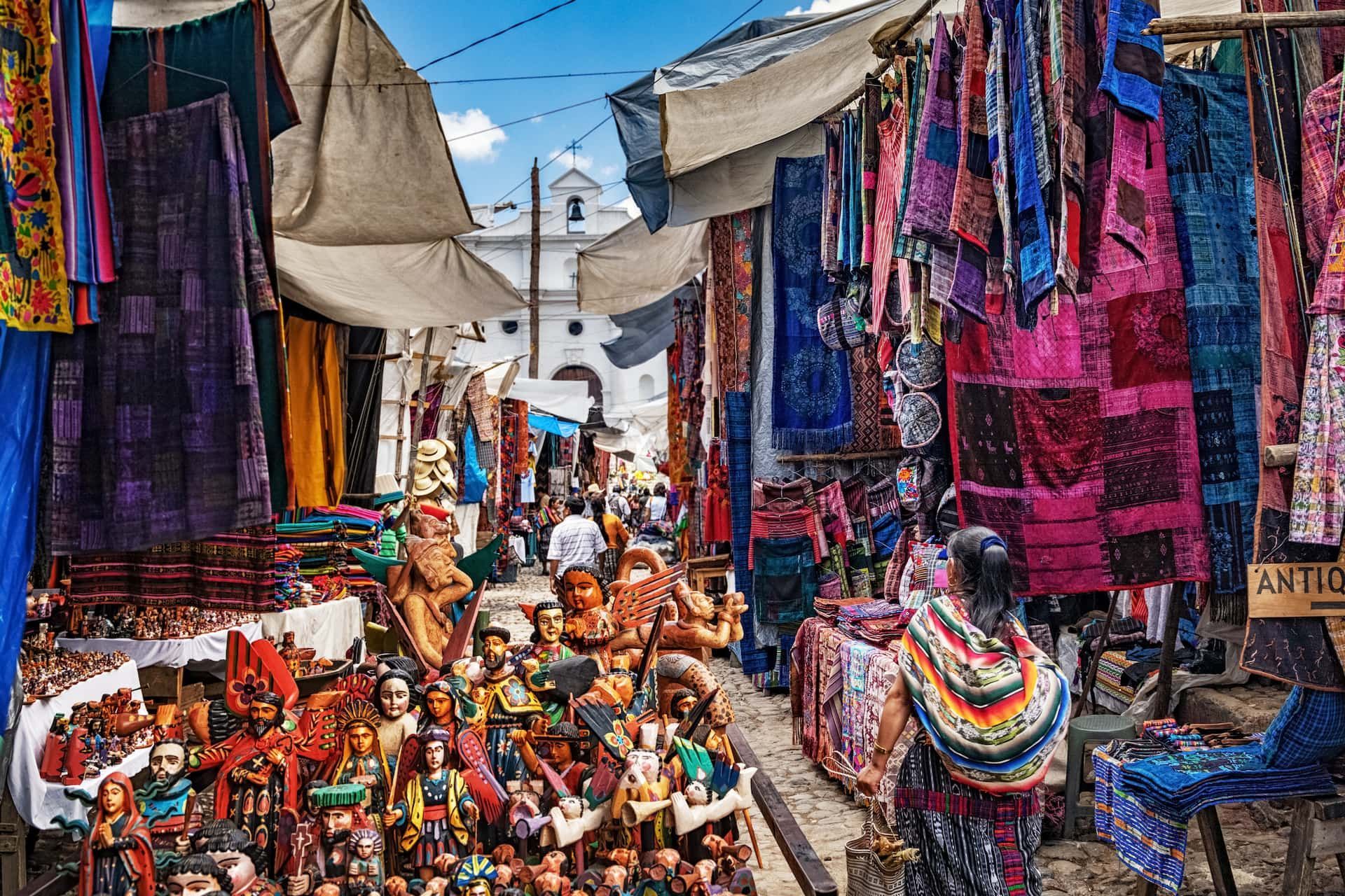 Un marché de rue animé, débordant de textiles colorés et à motifs et d'artisanat en bois sculpté, sous un ciel ensoleillé.