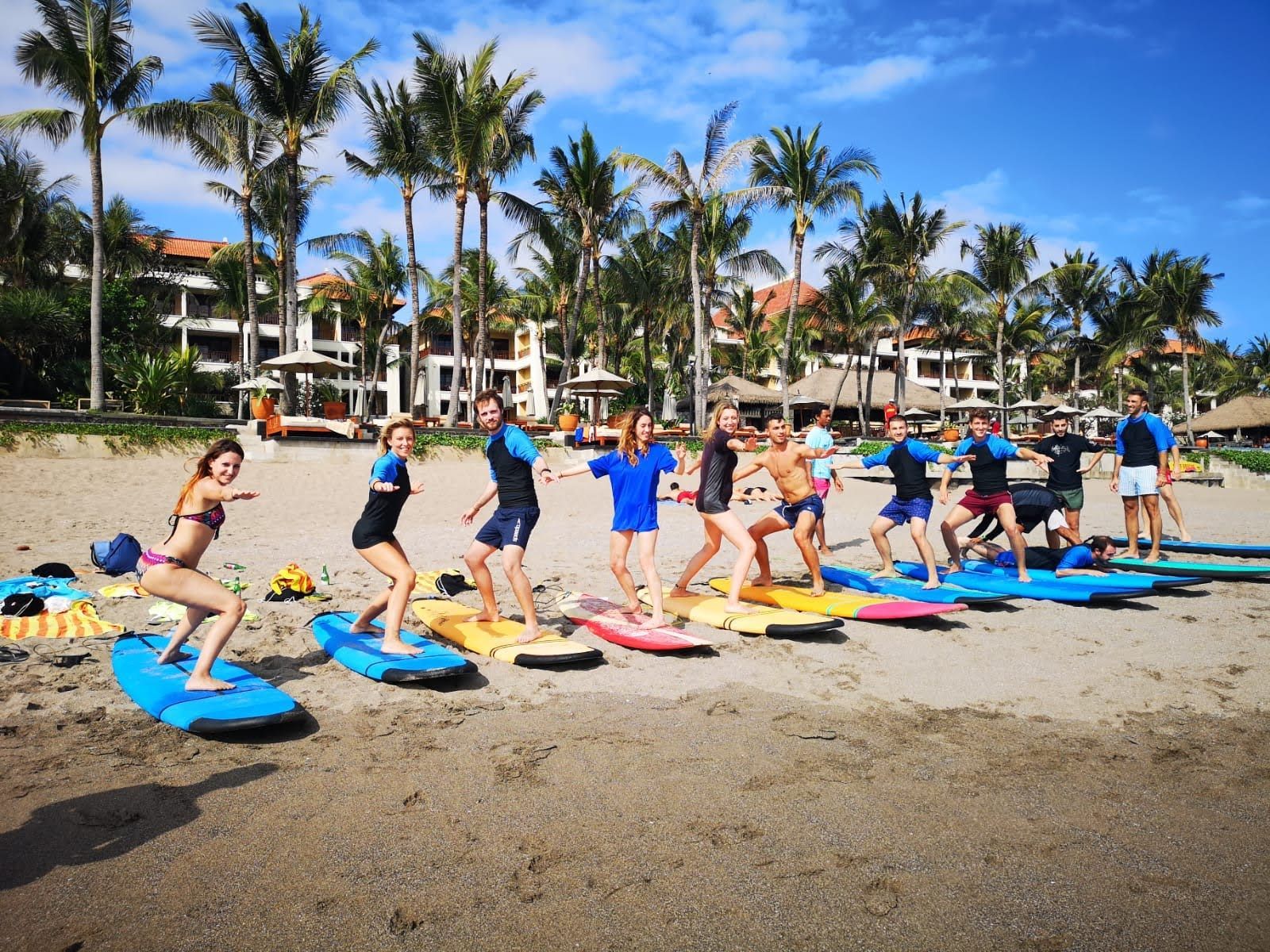 Un groupe WeRoad sur la route, prenant un cours de surf sur une plage de sable, s'entraînant sur des planches de surf colorées sous des palmiers.