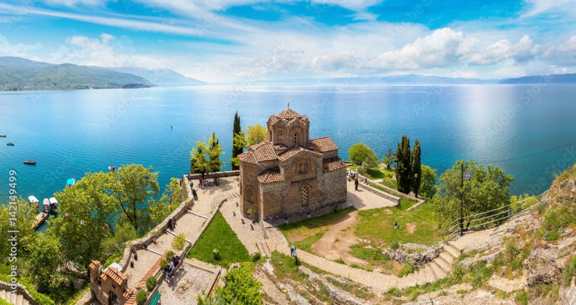 Una vista panorámica de una antigua iglesia de piedra en un acantilado con vistas a un vasto lago azul, con montañas distantes bajo un cielo nublado.