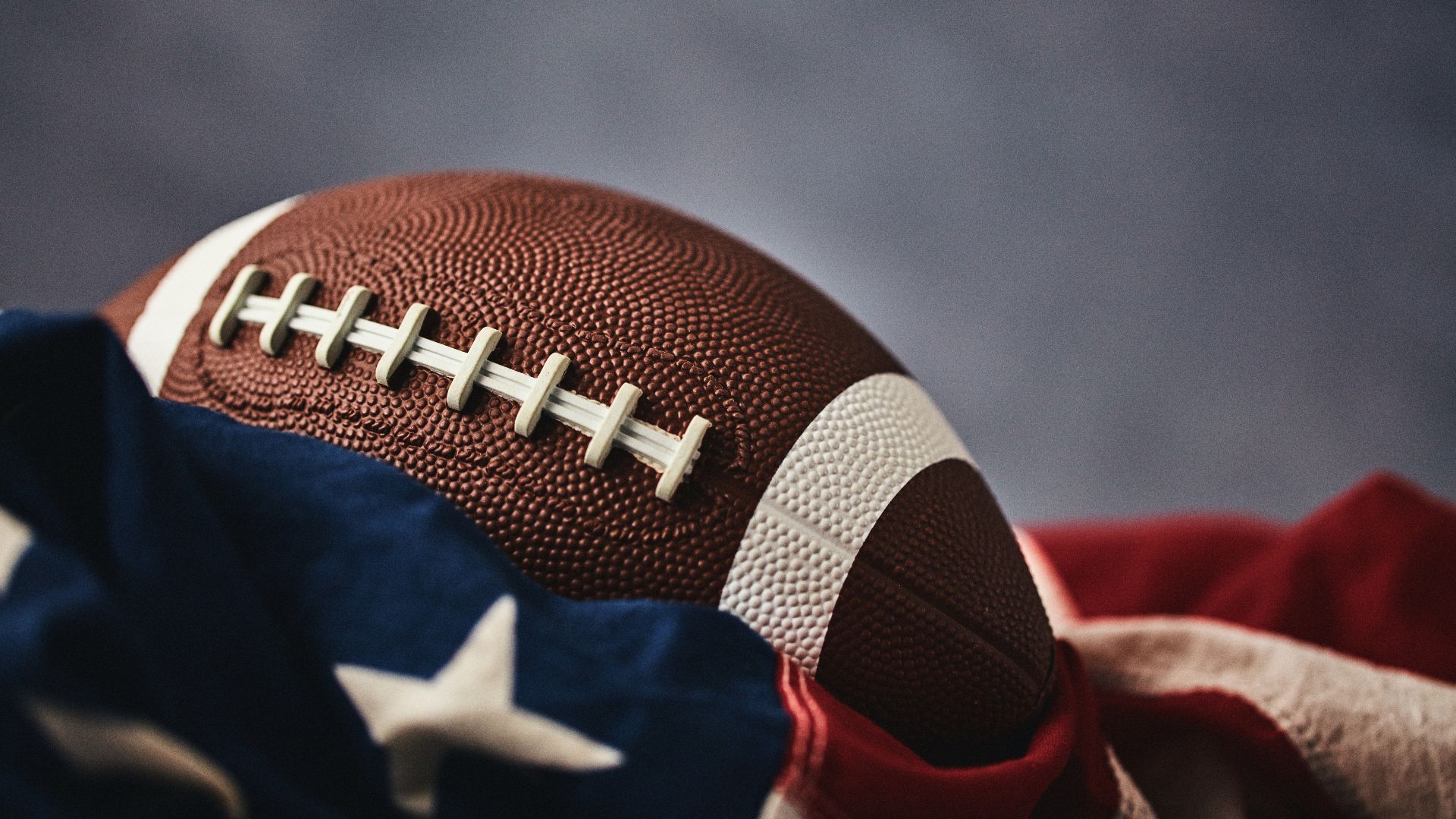 Primo piano di un pallone da football americano marrone con lacci bianchi adagiato su una bandiera americana drappeggiata.