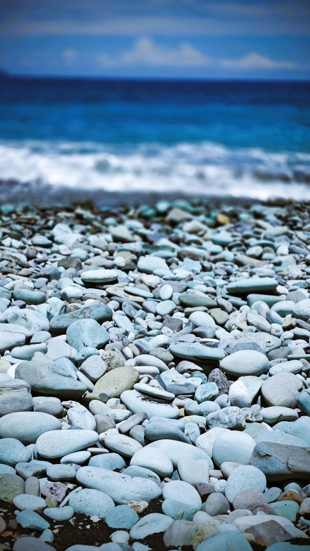 Nahaufnahme von glatten, hellblauen Kieselsteinen am Strand, mit dem tiefblauen Meer unscharf im Hintergrund.