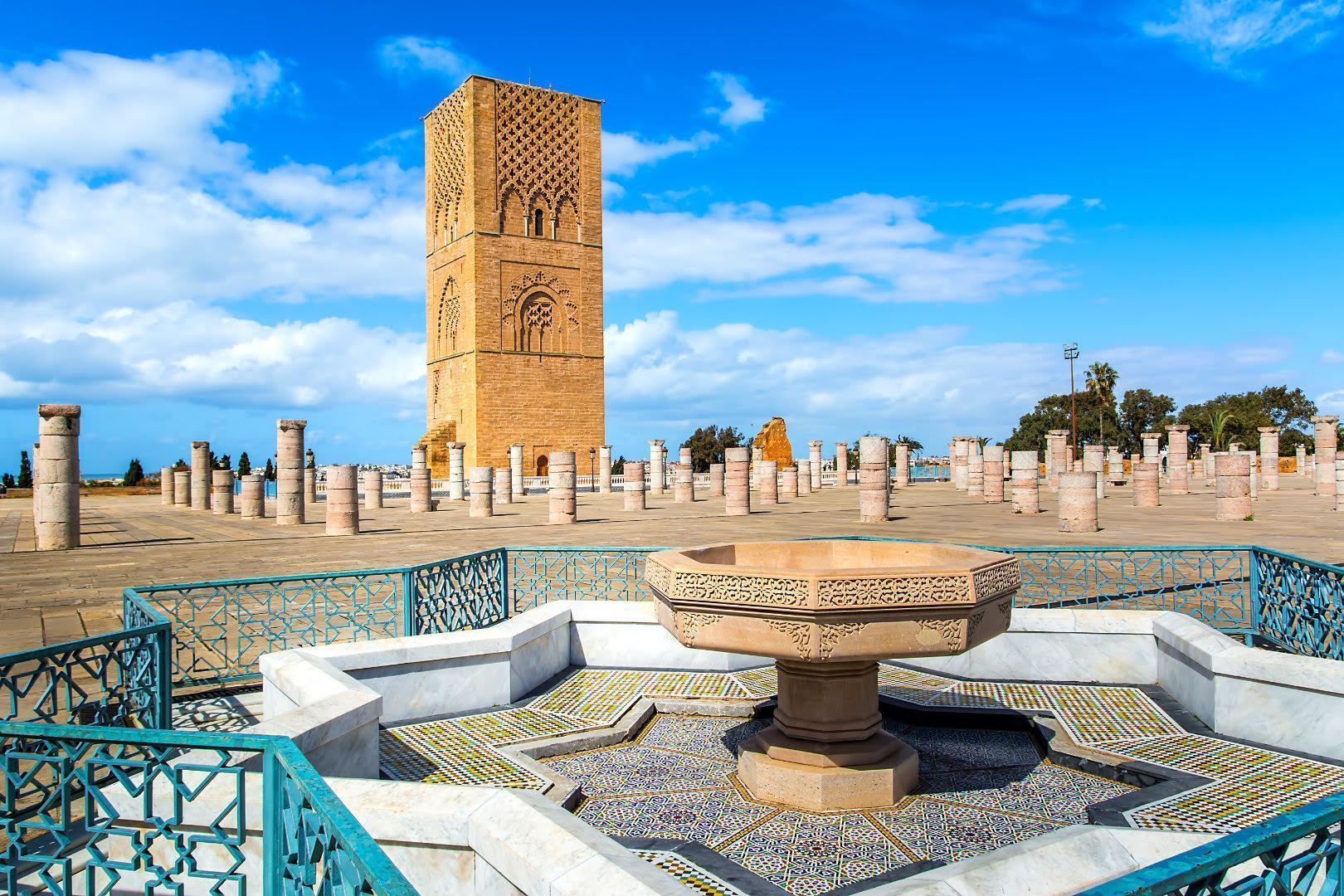 A tall, square, sand-colored stone tower stands behind rows of ancient columns in a plaza, with an ornate fountain in the foreground.