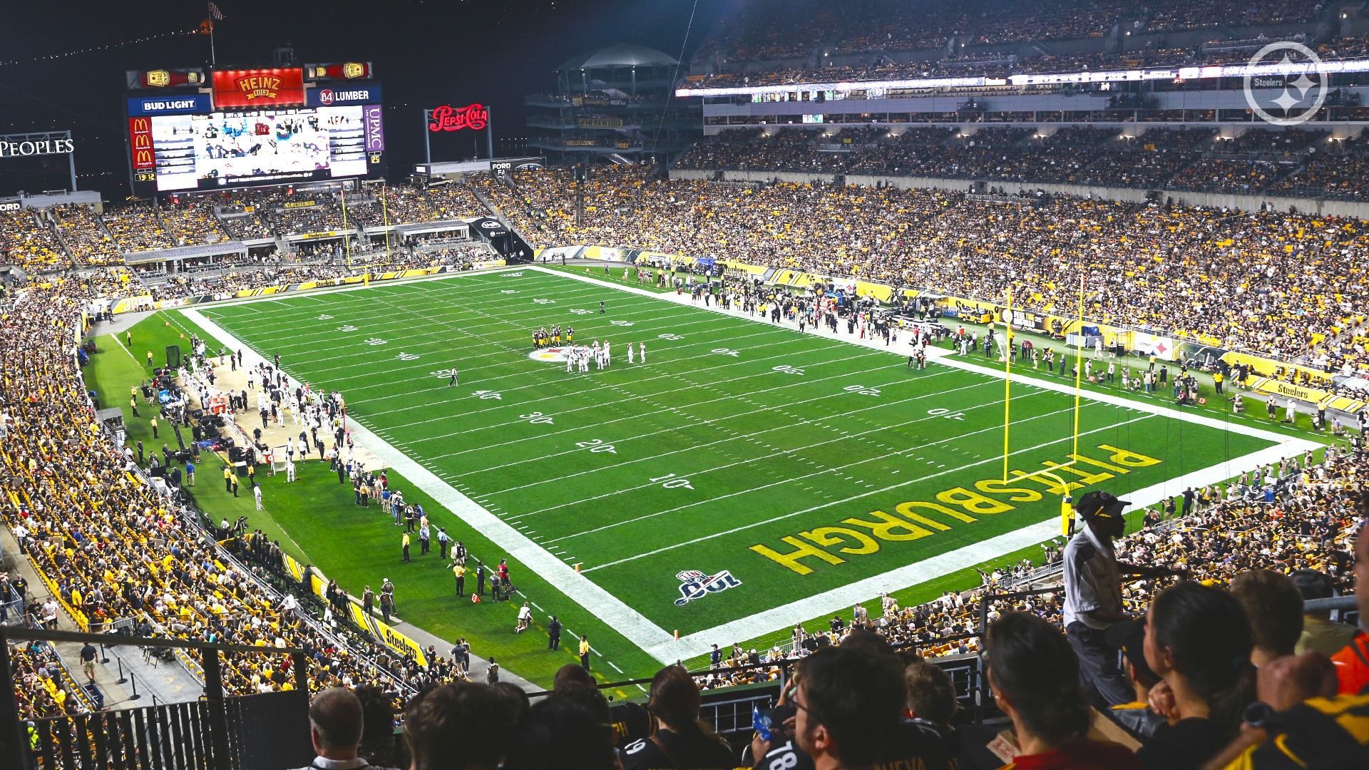 Una vista panoramica di uno stadio di football americano affollato durante una partita notturna, con 'Pittsburgh' dipinto nella endzone del campo.