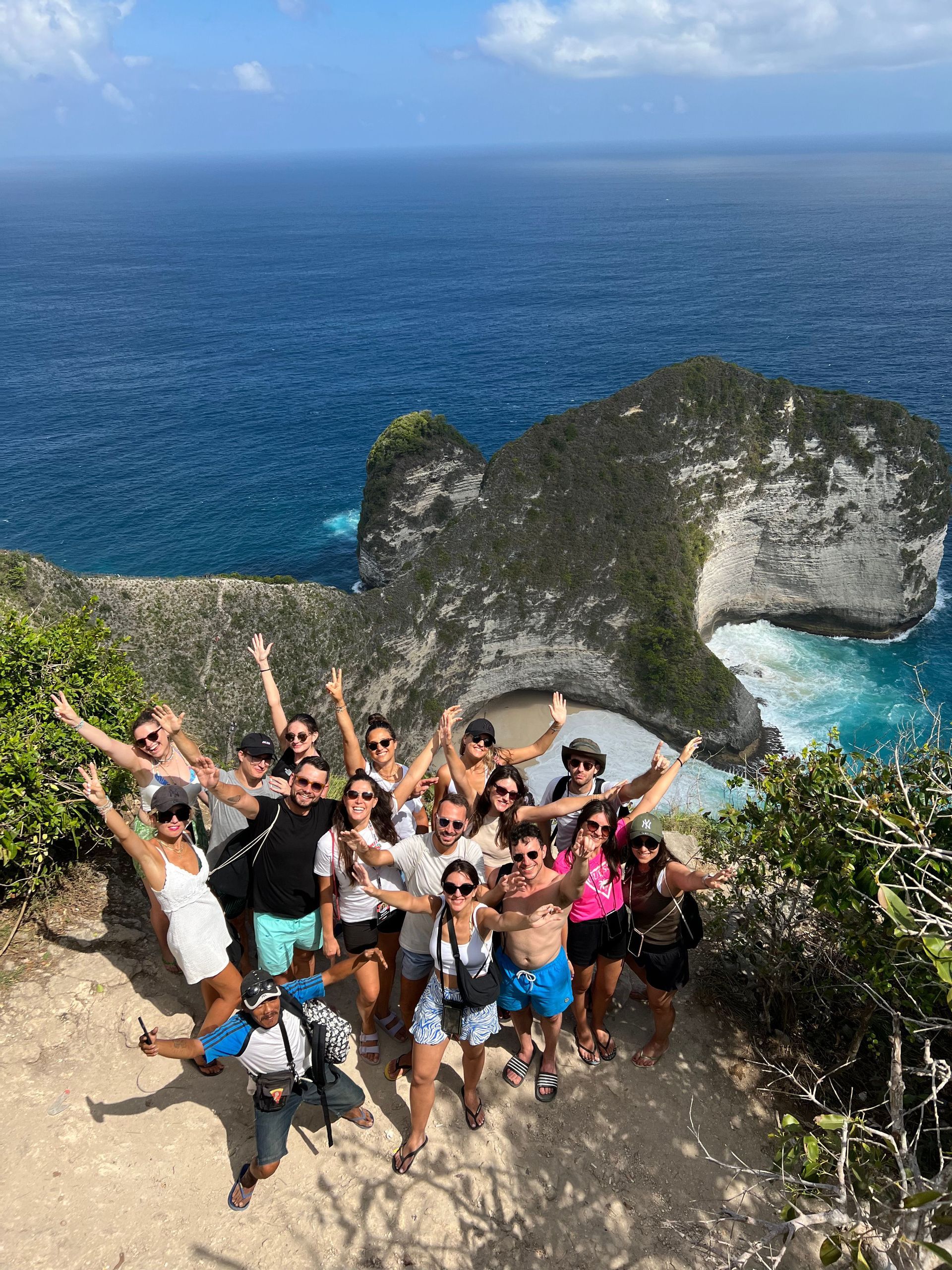 Un groupe WeRoad en voyage sourit et pose sur un point de vue de falaise avec une formation rocheuse spectaculaire et l'océan turquoise en contrebas.