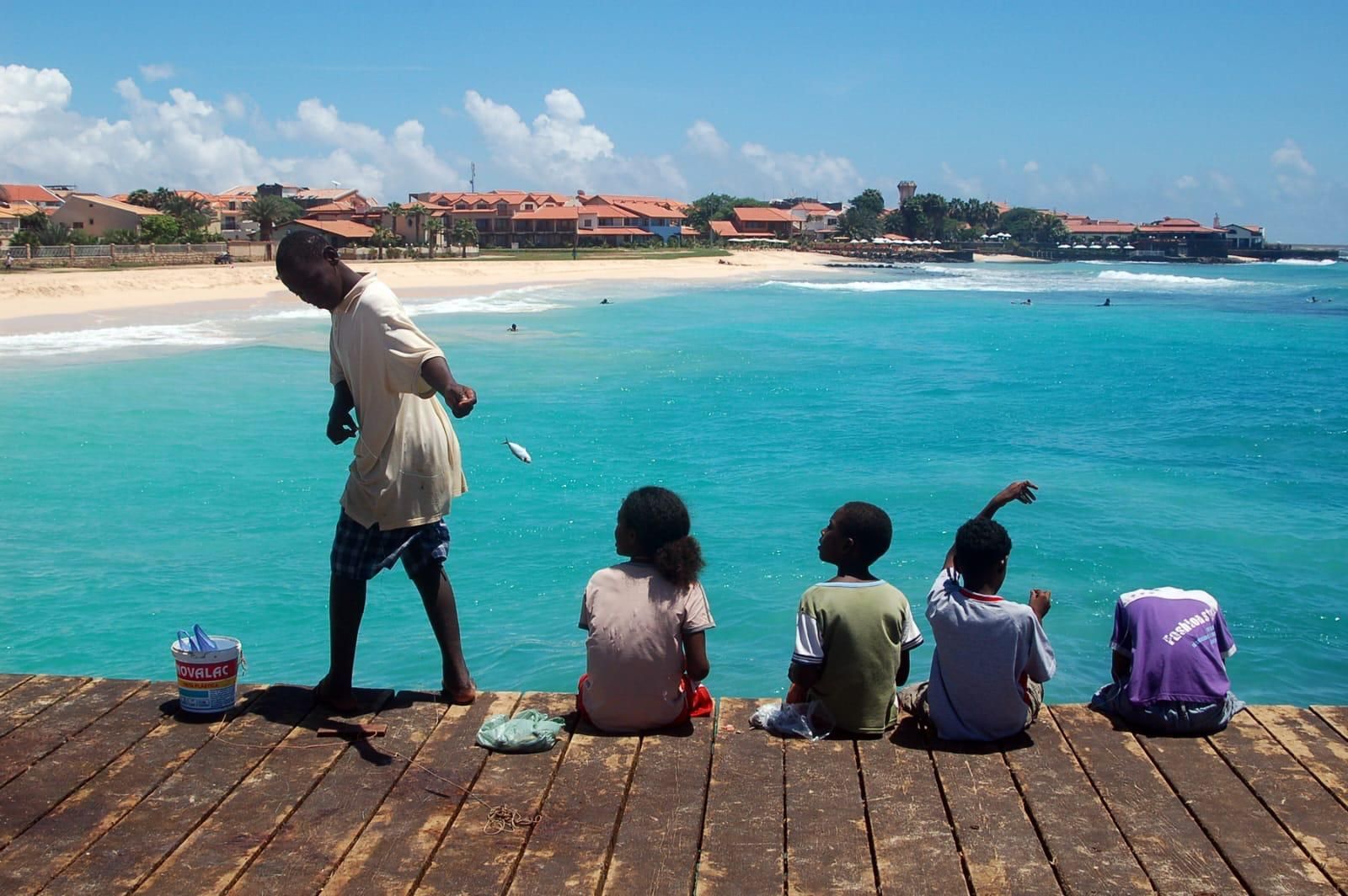 Un ragazzo su un pontile di legno accanto a tre bambini seduti, con un piccolo pesce sospeso sull'oceano turchese.