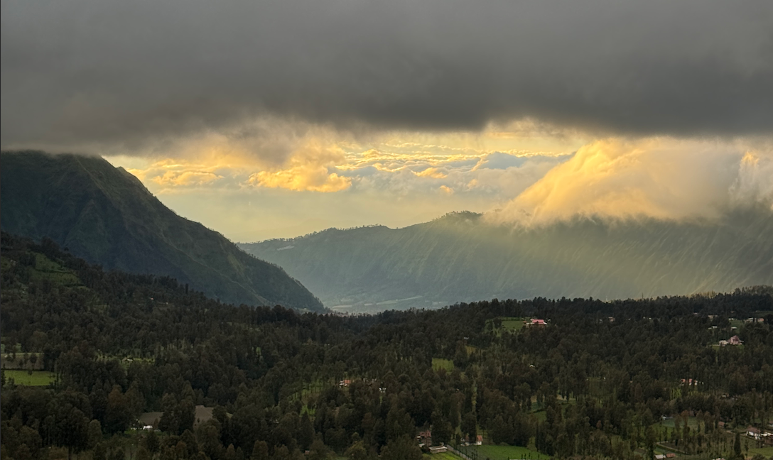 Ein üppiges grünes Tal und bewaldete Berge unter einem dunklen, bewölkten Himmel mit durchbrechendem goldenem Sonnenlicht.