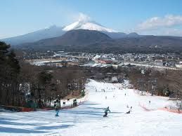 Persone che sciano e fanno snowboard su un'ampia pista con una città e un grande vulcano innevato sullo sfondo.