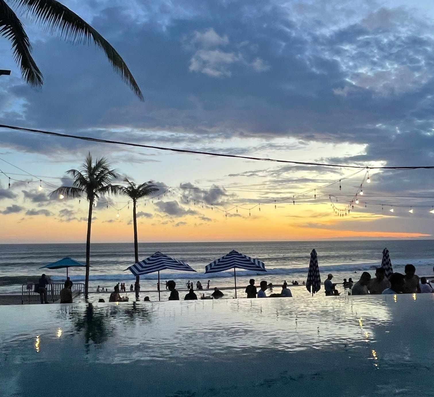 Eine WeRoad-Gruppe beobachtet den Sonnenuntergang von einem Infinity-Pool aus mit Blick auf einen Strand mit Palmen und Lichterketten.