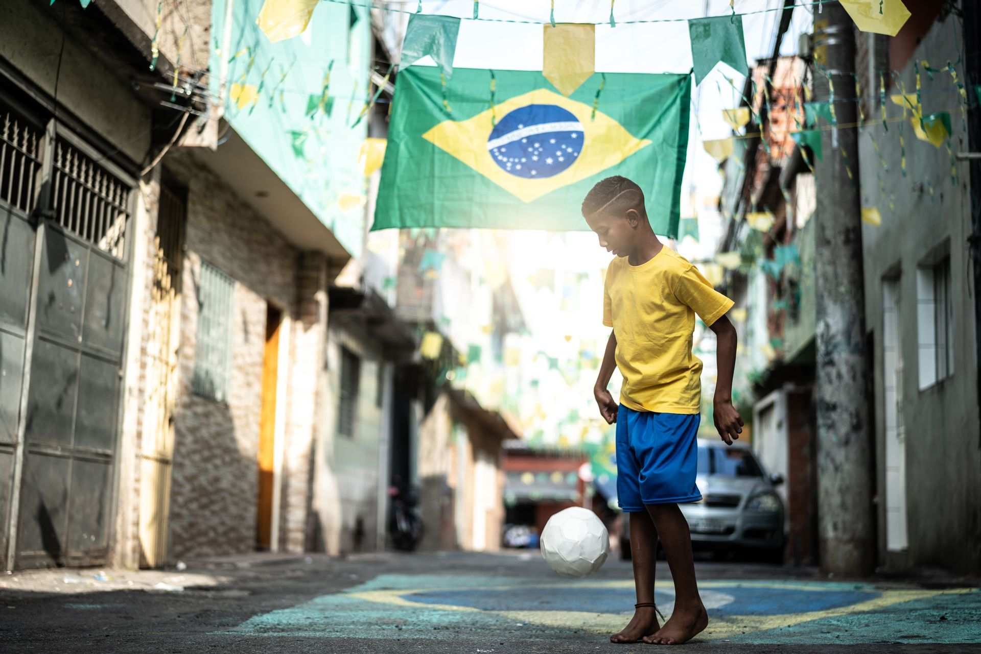 Un ragazzo a piedi nudi con una maglietta gialla gioca con un pallone da calcio su una strada decorata con bandiere brasiliane.