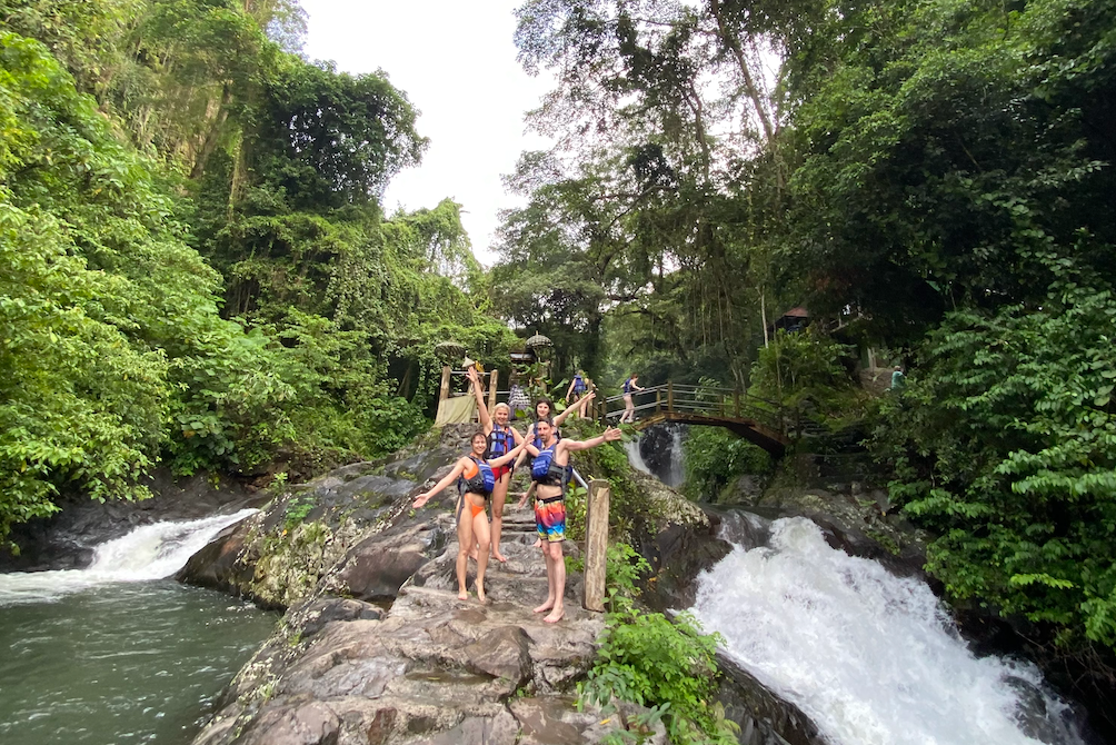Eine WeRoad-Gruppenreise posiert mit offenen Armen auf Felsen neben einem rauschenden Wasserfall in einem dichten Dschungel.