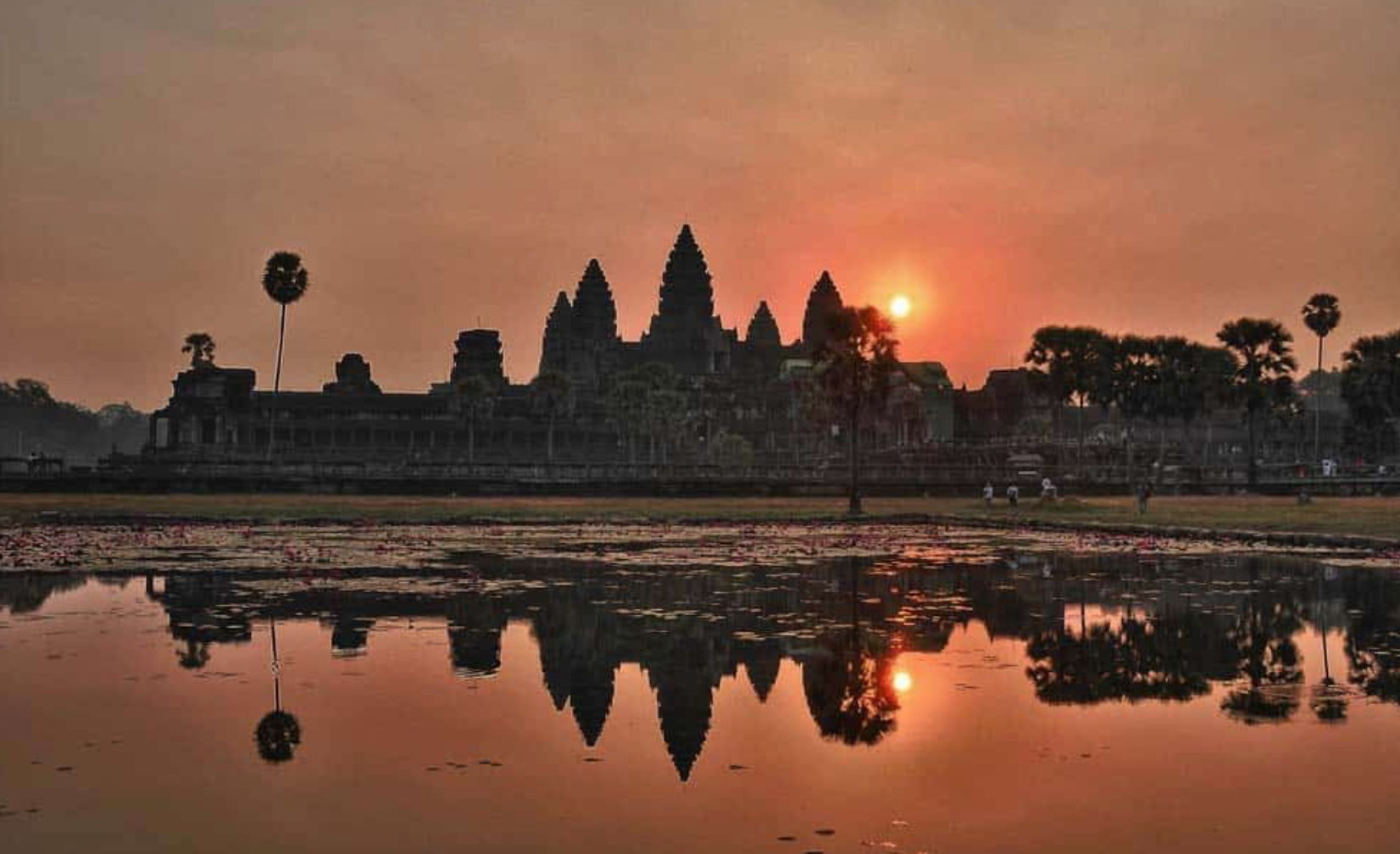 Die Silhouette eines großen alten Tempels bei Sonnenaufgang, wobei sich seine Türme und Form in einem stillen Teich im Vordergrund spiegeln.