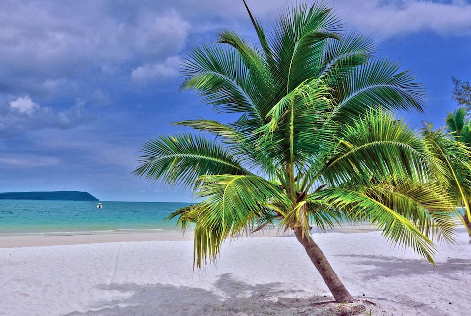 Eine Palme lehnt sich über einen weißen Sandstrand neben türkisfarbenem Wasser unter blauem Himmel mit vereinzelten Wolken.