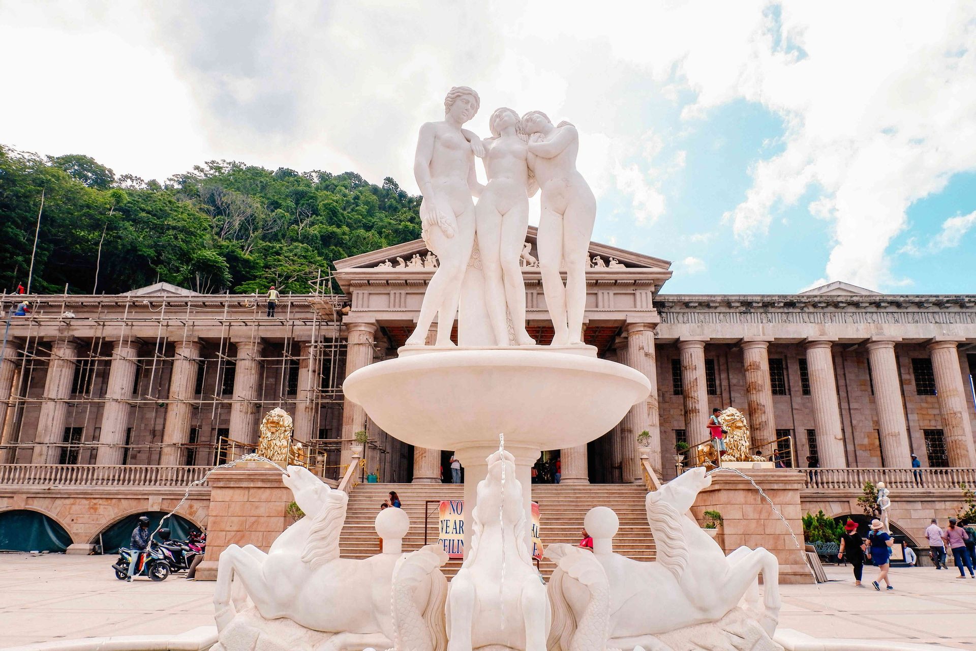 Una gran fuente con estatuas clásicas blancas se alza frente a un edificio columnado con andamios, teniendo como telón de fondo una colina boscosa.