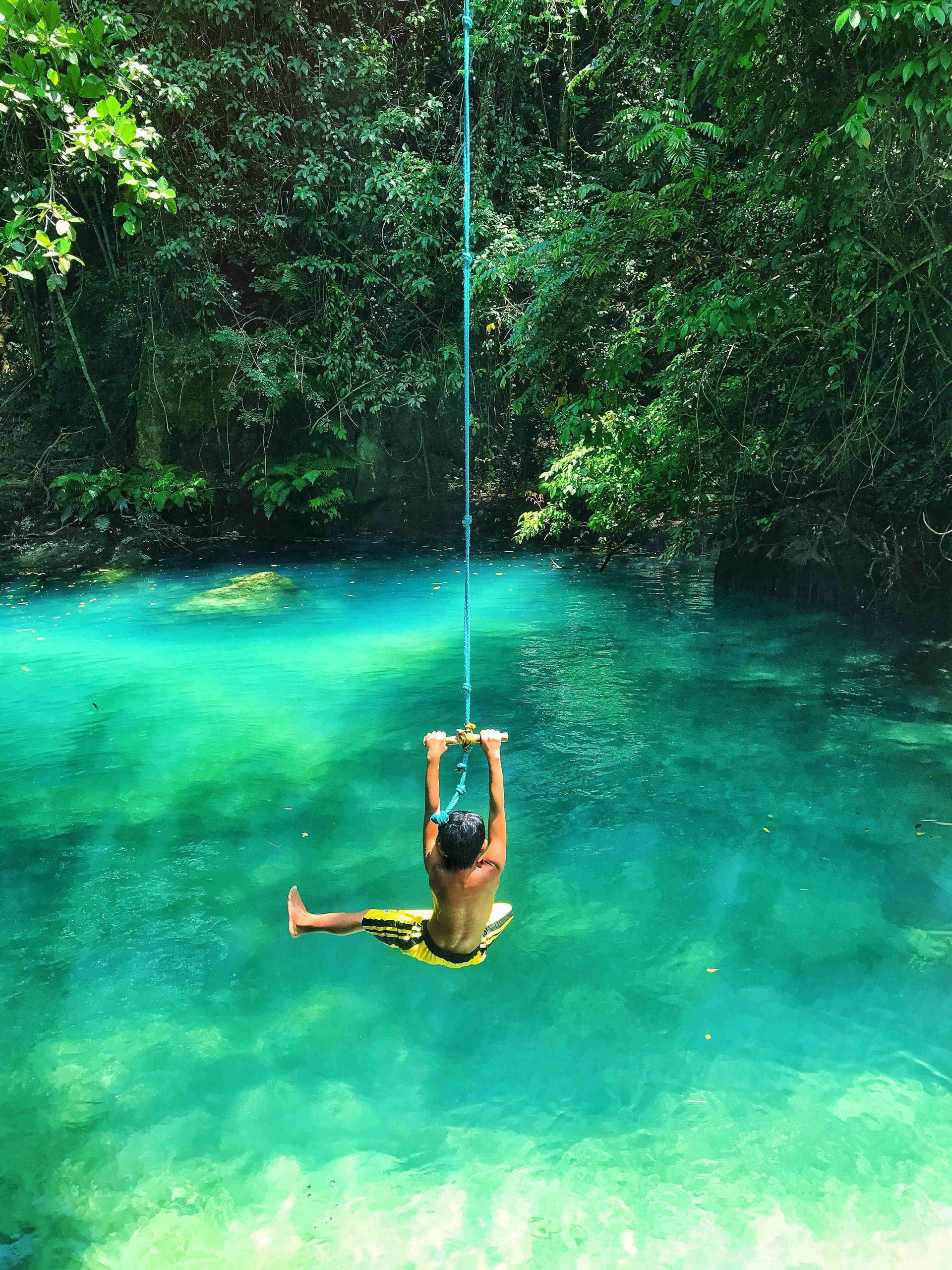 Una persona vista de espaldas en un columpio de cuerda se desliza sobre agua turquesa, con un denso bosque verde de fondo.