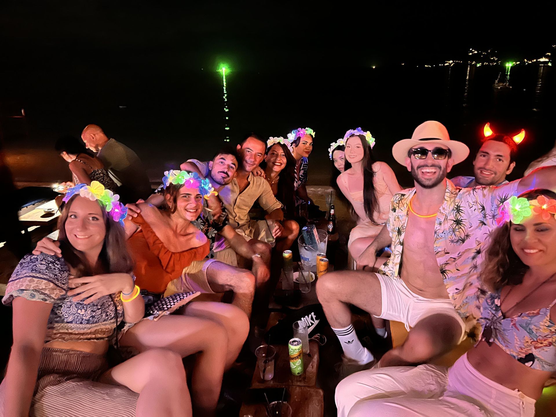Un grupo de WeRoad posa para una foto durante una fiesta nocturna en la playa, luciendo festivas coronas de flores luminosas.