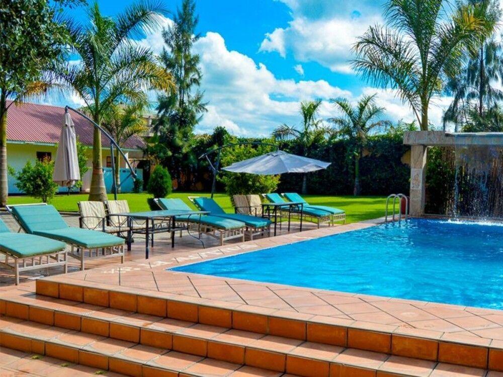 A swimming pool with a waterfall feature, surrounded by empty lounge chairs, palm trees, and a green lawn under a blue sky with clouds.
