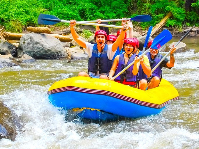 A WeRoad group trip smiles and raises their paddles while whitewater rafting on a river.