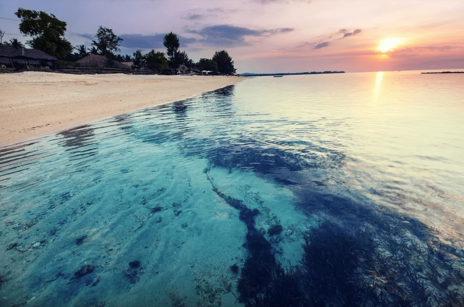 Clear turquoise water with a visible seabed of sand and seaweed laps against a sandy beach at sunset.