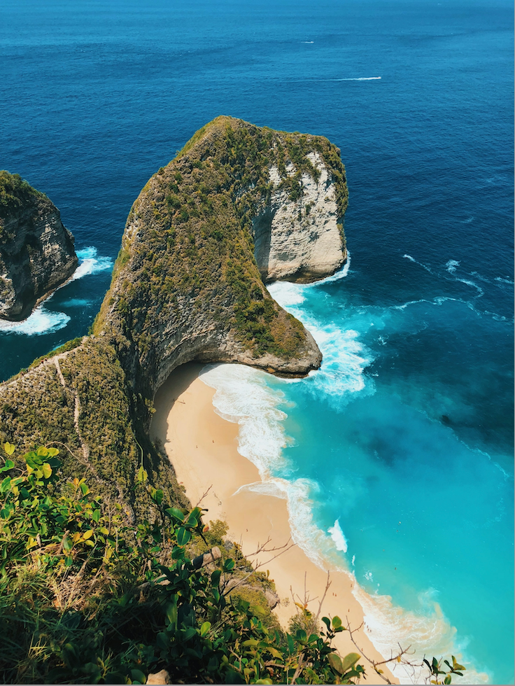 A high-angle view of a large, green-covered cliff formation curving over a secluded sandy beach and turquoise ocean.