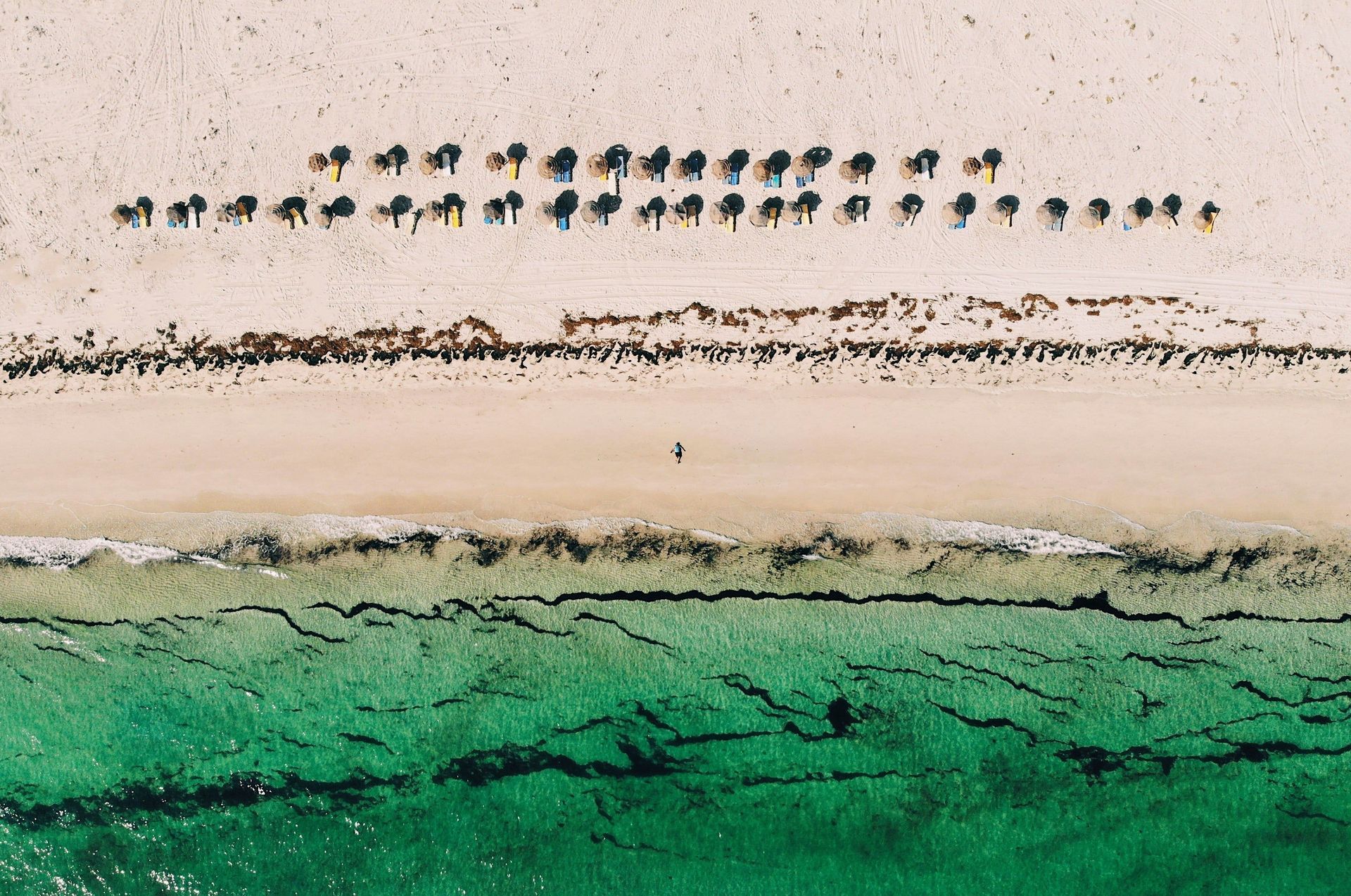Vue aérienne d'une personne marchant sur une plage de sable blanc, à côté d'une eau turquoise et de rangées de parasols en paille.
