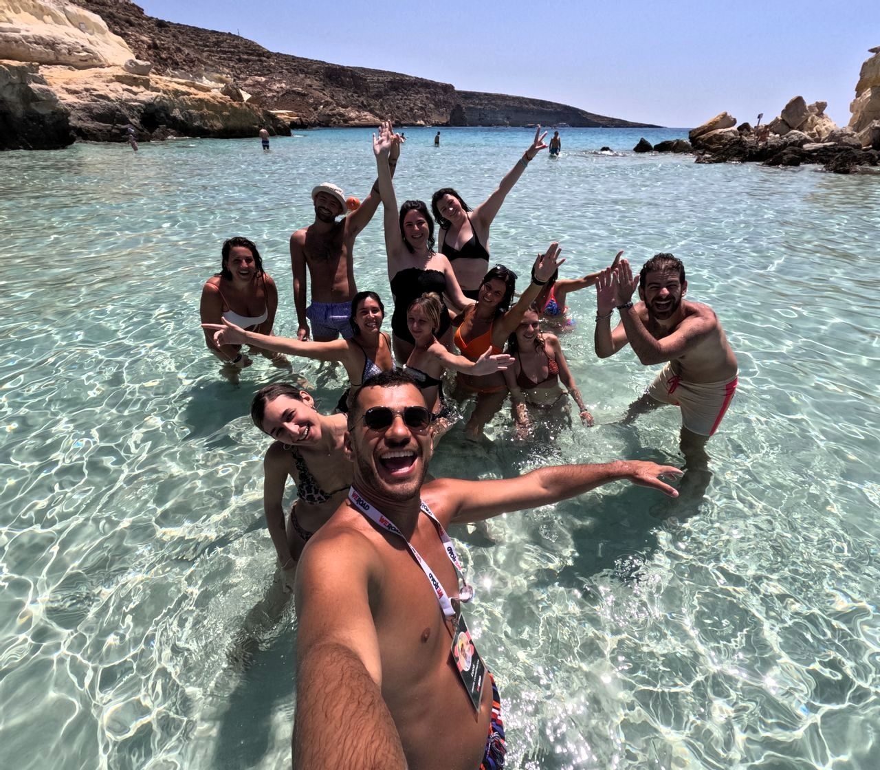 Un gruppo WeRoad sorride e posa per un selfie di gruppo, immerso nelle acque limpide e poco profonde di una costa rocciosa.