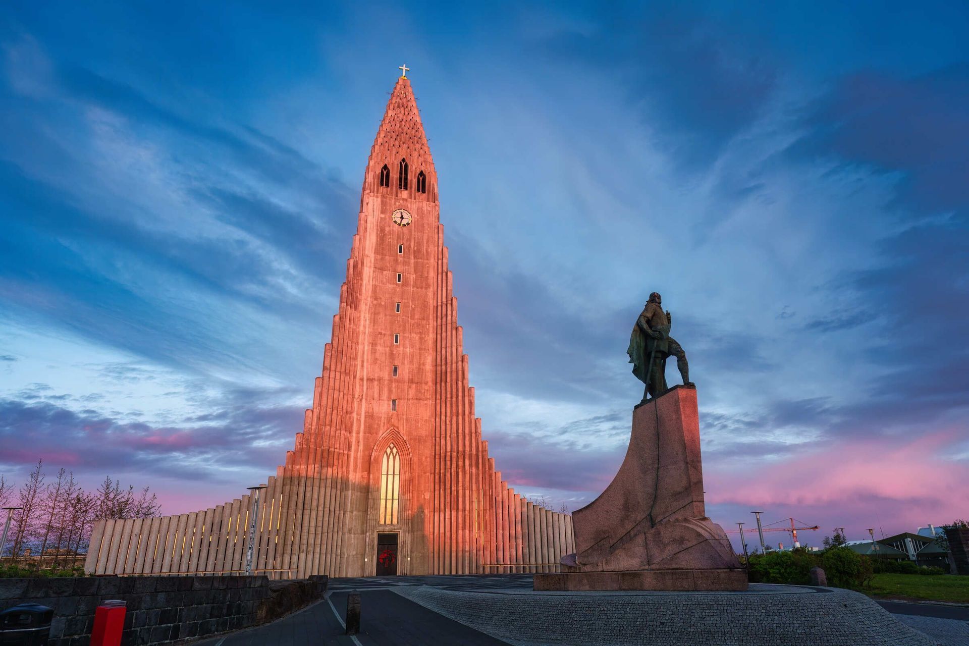 Eine hohe, moderne Kirche und eine Statue auf einem Sockel werden vom warmen Schein des Sonnenuntergangs vor einem dramatischen, bewölkten Himmel beleuchtet.