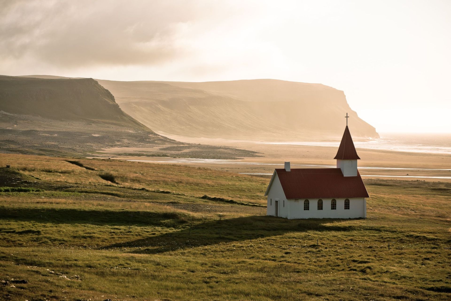 Eine weiße Kirche mit rotem Dach und Turm steht auf einem grasbewachsenen Feld mit dunstigen Klippen und einer Küste im Hintergrund.