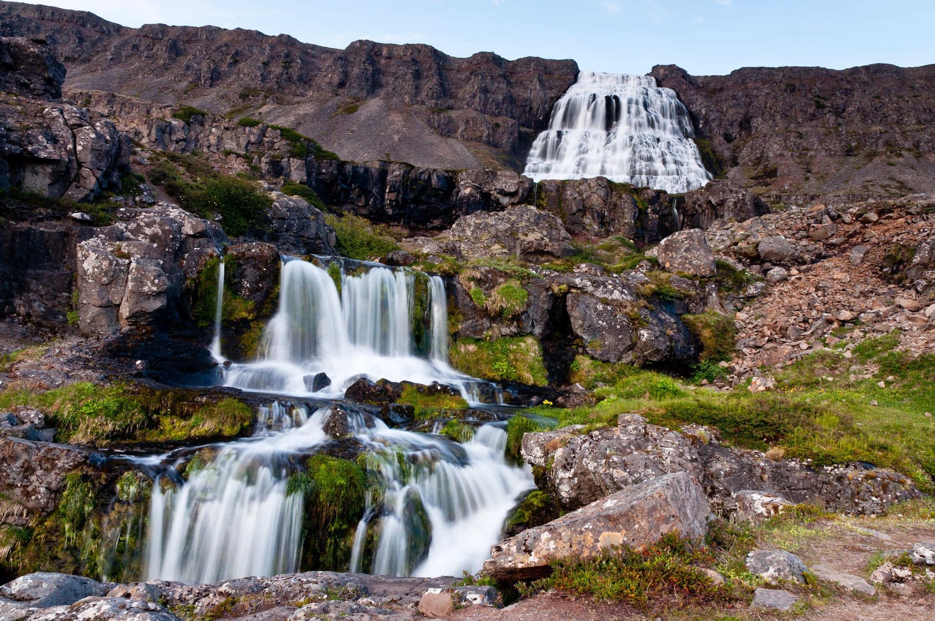 Ein mehrstufiger Wasserfall stürzt einen felsigen Berg hinab, wobei kleinere Wasserfälle im Vordergrund über moosbewachsene Felsen fließen.