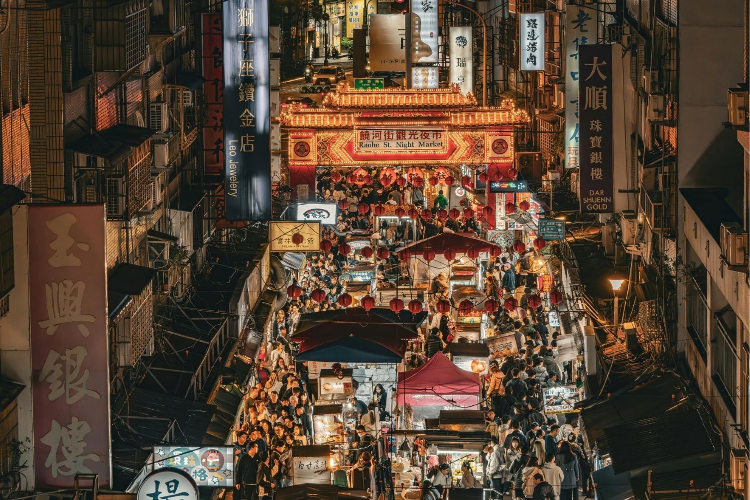 Eine Vogelperspektive auf einen belebten asiatischen Nachtmarkt, auf dem Menschen zwischen Ständen unter beleuchteten Schildern und roten Laternen umhergehen.