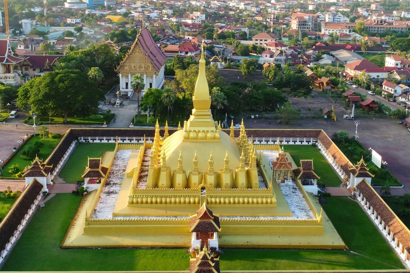 Eine Luftaufnahme einer großen, goldenen Stupa mit mehreren Türmen, umschlossen von einem ummauerten Hof, mit einer Stadt im Hintergrund.