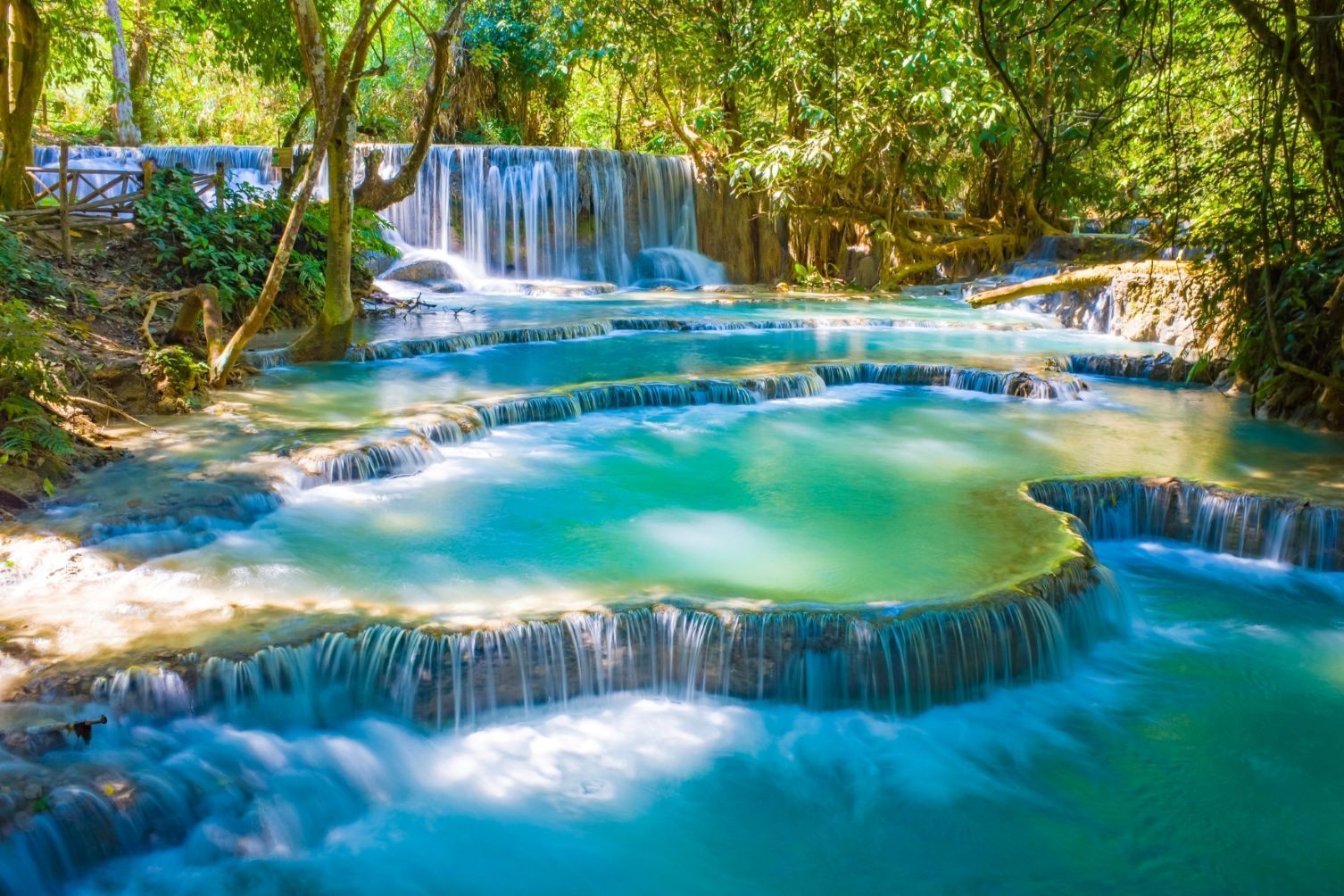Ein breiter, mehrstufiger Wasserfall fließt in mehrere türkisfarbene Becken in einem sonnendurchfluteten grünen Wald.
