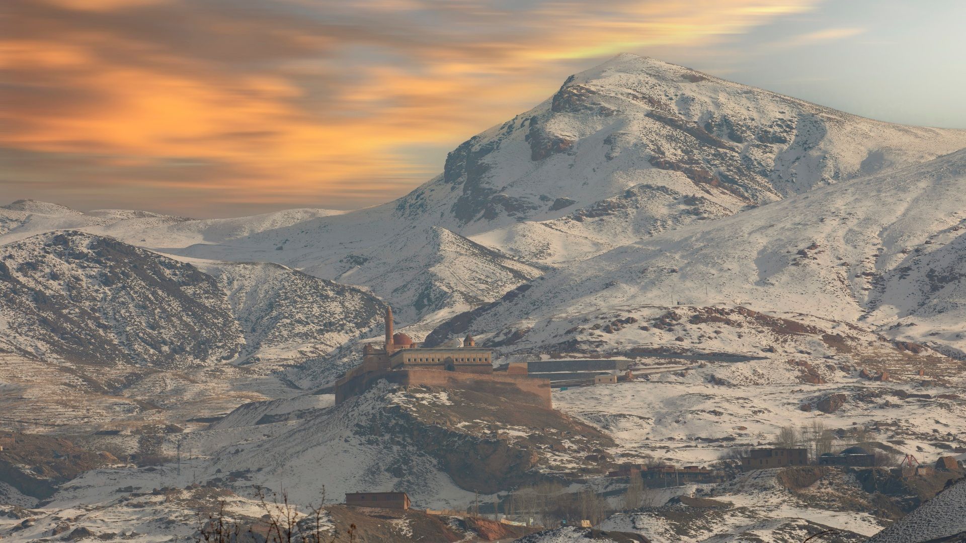 Un palazzo storico e un minareto si ergono su una collina circondata da vaste montagne innevate sotto un cielo colorato al tramonto.