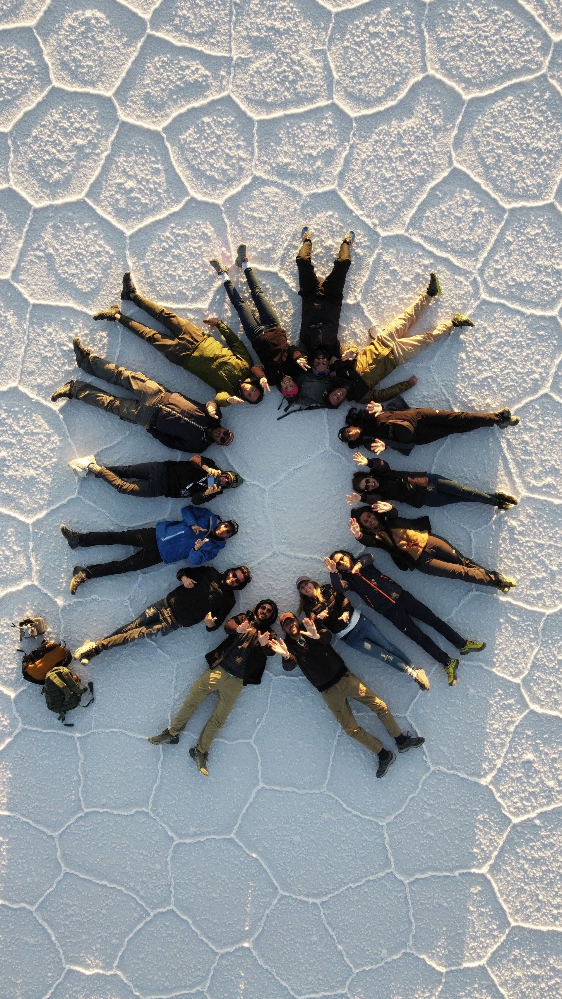 Vista aerea di un gruppo WeRoad sdraiato in cerchio su una vasta piana di sale con crepe poligonali.